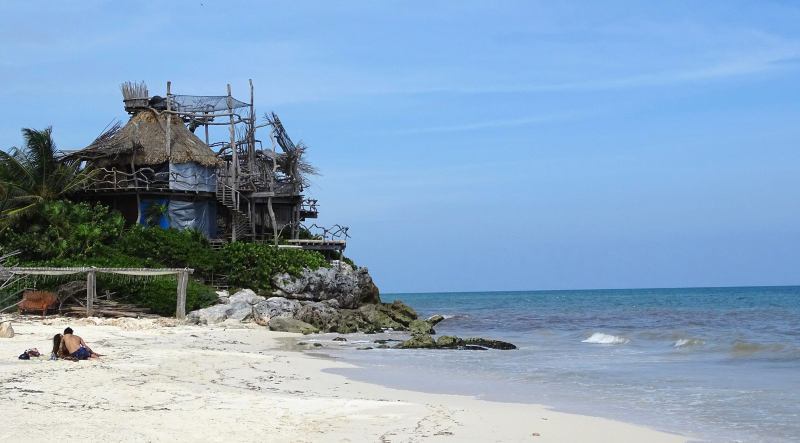 The beach at Tulum with a driftwood and thatch building on the rocks above it
